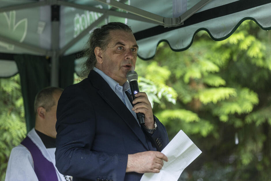 Na zdjęciu mężczyzna w ciemnej marynarce i jasnoniebieskiej koszuli przemawia do mikrofonu, trzymając w ręce kartkę z tekstem. W tle widoczny jest duchowny oraz zielone gałęzie, co sugeruje, że scena ma miejsce podczas uroczystości plenerowej, prawdopodobnie pogrzebowej. 