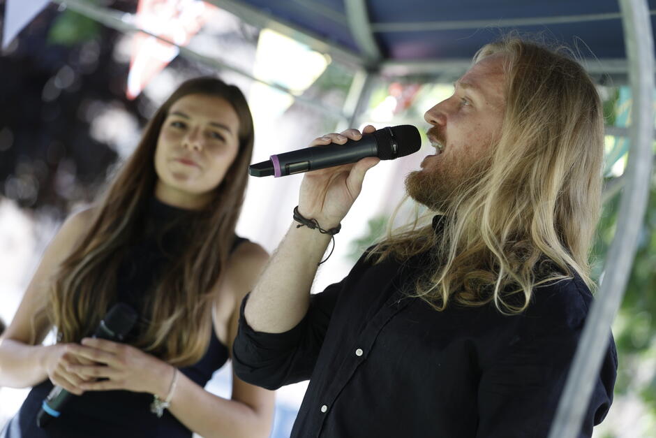 Dwójka wokalistów wykonuje utwór podczas występu na scenie plenerowej — mężczyzna z długimi włosami śpiewa do mikrofonu, a kobieta towarzyszy mu z mikrofonem w dłoniach. Kameralna scena pod namiotem i letnia atmosfera podkreślają swobodny charakter koncertu.