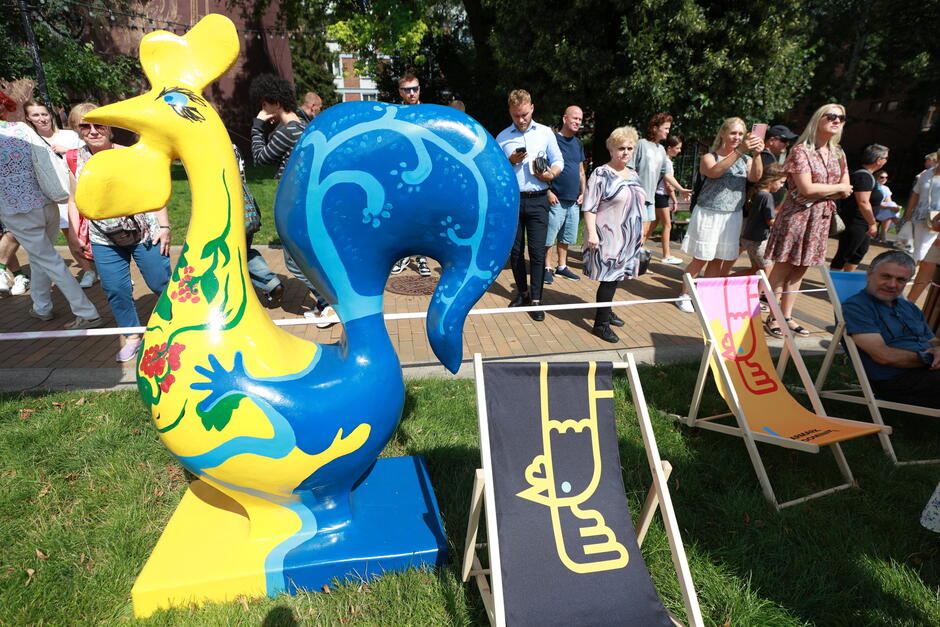 Kolorowa rzeźba koguta — symbolu Jarmarku św. Dominika — stoi na trawie wśród przechodniów i odwiedzających jarmark. W tle widać spacerujących ludzi oraz leżaki z charakterystycznym motywem graficznym, tworzące przyjazną przestrzeń dla uczestników wydarzenia