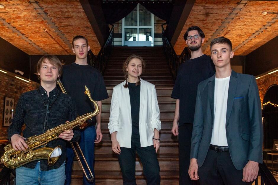 Członkowie Hania Derej Quintet pozują razem na tle eleganckiego wnętrza z drewnianymi schodami — muzycy trzymają swoje instrumenty i uśmiechają się do obiektywu. Zdjęcie oddaje młodzieńczy charakter zespołu i ich jazzową tożsamość.