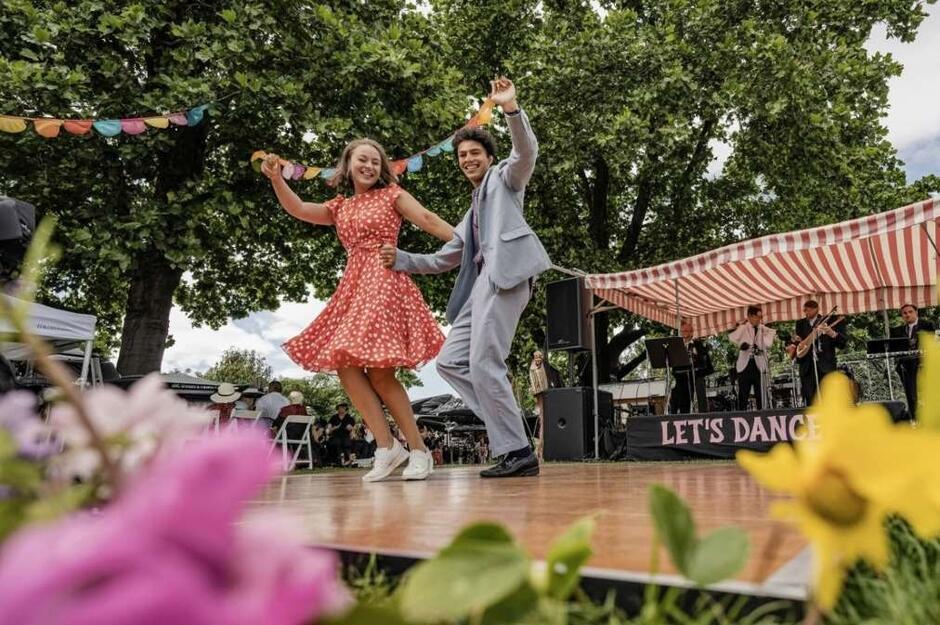 Na zdjęciu widać parę młodych tancerzy tańczących na drewnianym parkiecie na świeżym powietrzu, obok sceny z grającym zespołem pod biało-czerwonym namiotem z napisem „LET’S DANCE”. W tle widać publiczność, dekoracje w postaci kolorowych chorągiewek i drzewa, co nadaje wydarzeniu festynowy, radosny charakter