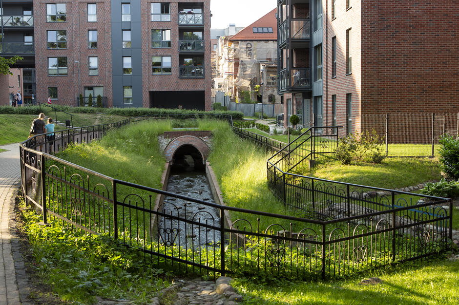 Na zdjęciu widoczny jest strumień płynący przez osiedle z nowoczesnymi budynkami z czerwonej cegły, częściowo zakryty ceglanym przepustem. Wzdłuż nurtu biegnie ścieżka spacerowa z ozdobnym ogrodzeniem, przy której stoją ludzie obserwujący otoczenie. 