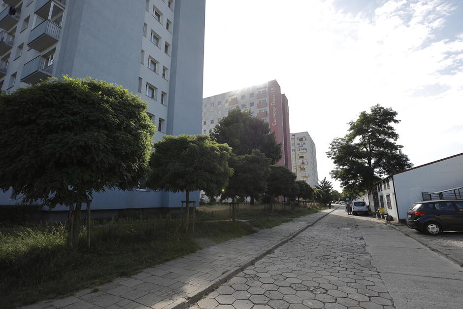 na zdjęciu pośrodku ulica o jezdni z kostki betonowej, po lewej trzy wysokie bloki mieszkalne, po prawej niższy budynek