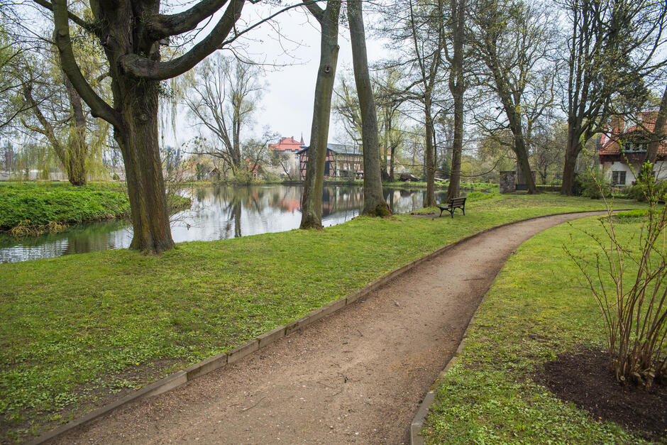 Zdjęcie przedstawia malowniczy park z alejką spacerową, otoczony drzewami i ławeczkami, biegnącą wzdłuż stawu z odbijającą się w wodzie zabudową o tradycyjnej architekturze. Miejsce emanuje spokojem i naturalnym pięknem, zachęcając do wypoczynku i spacerów wśród zieleni. 
