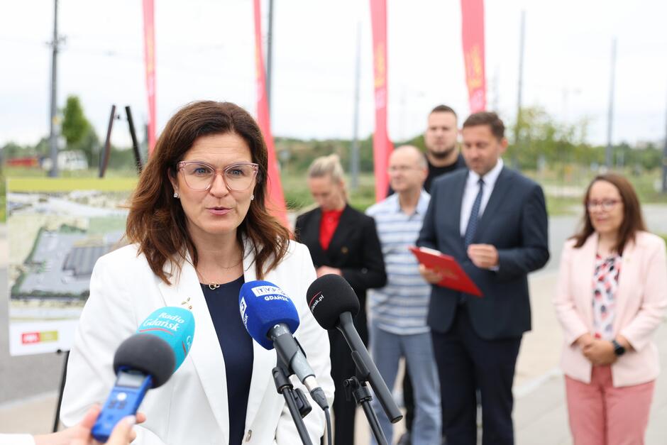 na zdjęciu kobieta w średnim wieku, w białej marynarce, mówi do dziennikarskich mikrofonów