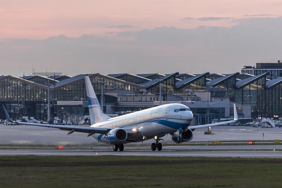Na zdjęciu widzimy samolot pasażerski linii Enter Air lądujący na pasie startowym przy świetle zachodzącego słońca. W tle znajduje się nowoczesny terminal lotniska z charakterystycznym przeszklonym dachem.
