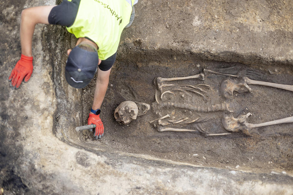 Widoczny pionowo, odkopany grób, a w nim szkielet. Wokół grobu widoczne z góry postaci i nogi z butami ekipy eksplorującej to miejsce 