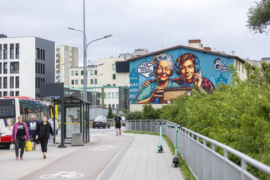 Przedstawia ulicę w Gdańsku z przystankiem autobusowym, pieszymi i ścieżką rowerową, a w tle znajduje się kolorowy mural na budynku. Mural przyciąga wzrok intensywnymi barwami i wizerunkiem starszej kobiety oraz młodego mężczyzny słuchających radia
