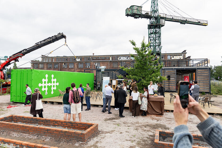 Na terenach poprzemysłowych stoją ludzie i obserwują jak dźwig wyładowuje z ciężarówki zielony kontener.