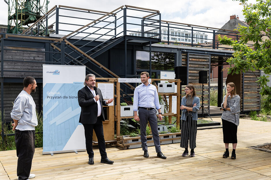 Pięć osób - trzech mężczyzn i dwie kobiety, stoją na drewnianym podeście przed obiektem zbudowanym z kontenerów. jeden mężczyzna przemawia do mikrofonu.