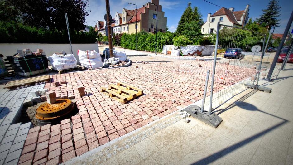 na zdjęciu fragment nowo układanej nawierzchni jezdni, w tle widać maszyny robotnicze, palety budowlane i ogrodzenie z siatki