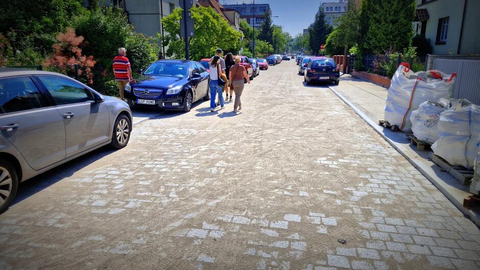 fragment wyremontowanej ulicy z nową nawierzchnią z kostki, idą po niej trzy dorosłe osoby, widać też sporo samochodów osobowych zaparkowanych po obu stronach ulicy