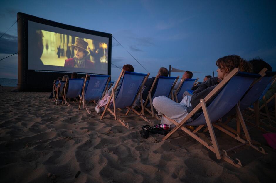 na zdjęciu leżaki ustawione na plaży, na nich siedzą ludzie, przed nimi wielki ekran na którym wyświetlany jest film