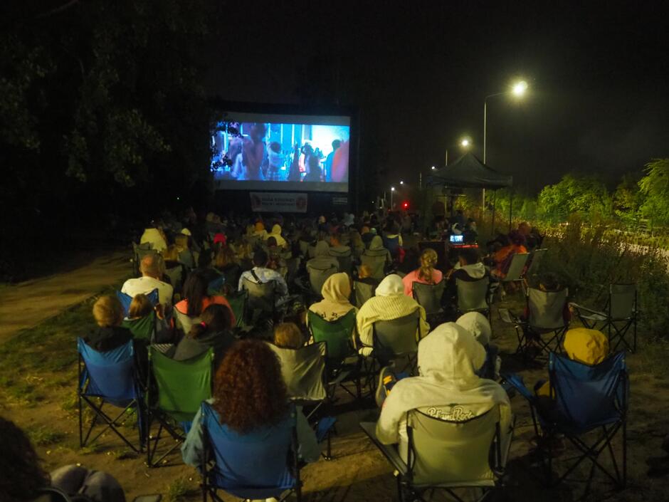 zdjęcie po zmroku, przed wielkim ekranem na którym wyświetlany jest film, siedzi na krzesełkach kilkadziesiąt osób