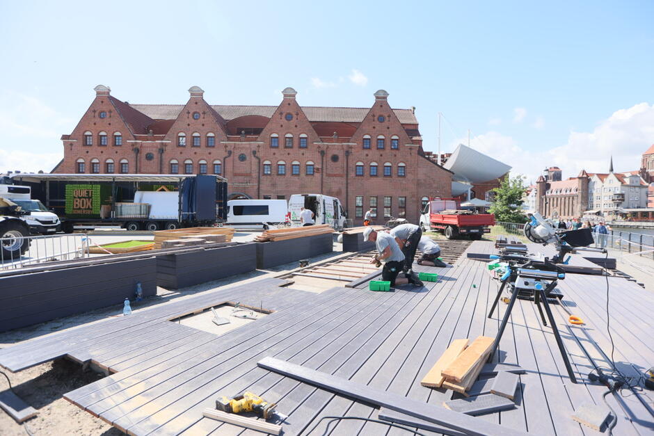 Na zdjęciu widać fragment nabrzeża, przy którym trwają prace budowlane – robotnicy pracują przy rozładunku materiałów obok zaparkowanego czerwonego auta dostawczego. Na wodzie obok nabrzeża płyną dwie osoby – jedna w kajaku, a druga w małej łodzi motorowej wyposażonej w koło ratunkowe.
