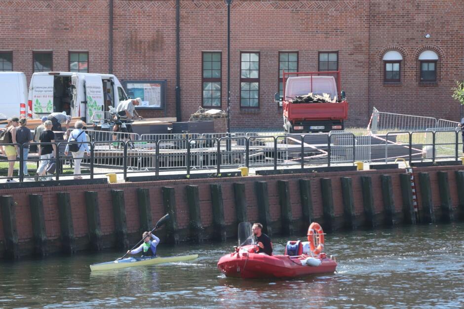 Na zdjęciu widać fragment nabrzeża, przy którym trwają prace budowlane – robotnicy pracują przy rozładunku materiałów obok zaparkowanego czerwonego auta dostawczego. Na wodzie obok nabrzeża płyną dwie osoby – jedna w kajaku, a druga w małej łodzi motorowej wyposażonej w koło ratunkowe.
