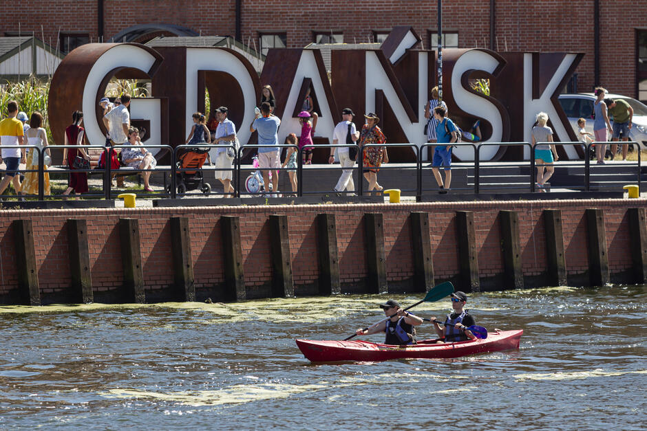 Na zdjęciu widoczna jest duża grupa turystów spacerujących nad Motławą w Gdańsku, w tle charakterystyczny napis „GDAŃSK”. Na pierwszym planie dwie osoby płyną czerwonym kajakiem po rzece, ciesząc się słoneczną pogodą.
