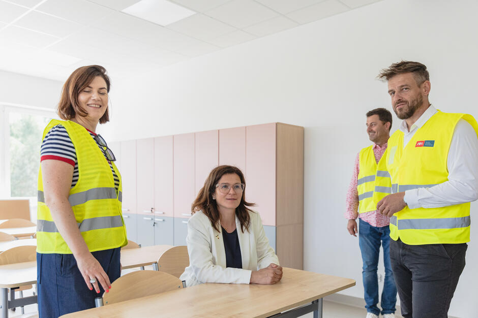 na zdjęciu kobieta w średnim wieku siedzi w szkolnej ławce, obok niej stoi dorosła kobieta i dwóch mężczyzn w średnim wieku