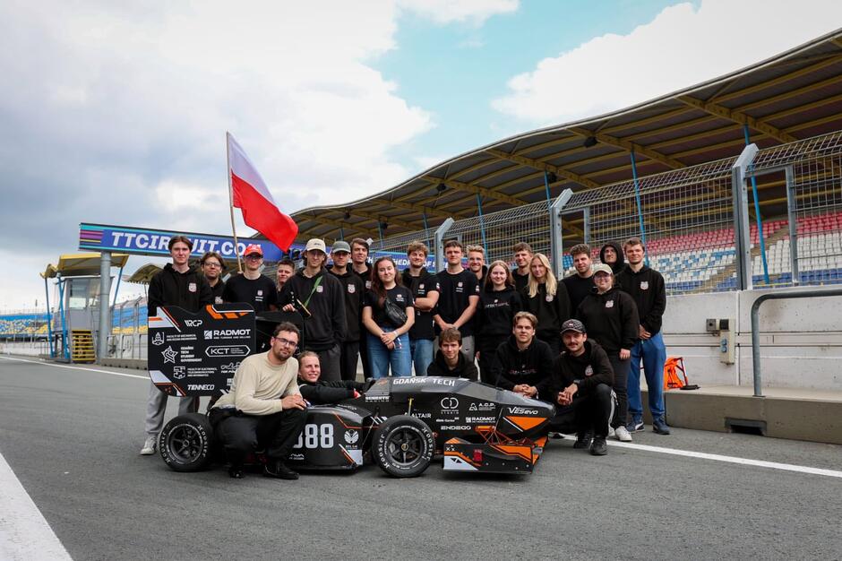 Kilkanaście osób stoi wokół bolidu wyścigowego na torze wyścigowym. W bolidzie siedzi jeden mężczyzna.