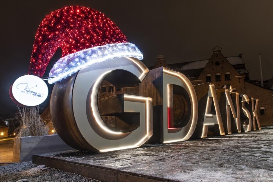 Na zdjęciu widnieje podświetlany napis GDAŃSK  ozdobiony dużą, świecącą czapką Mikołaja, co nadaje mu świąteczny charakter. Obok znajduje się logo Portu Lotniczego im. Lecha Wałęsy w Gdańsku, co sugeruje, że instalacja znajduje się w pobliżu lotniska lub jest z nim związana. 