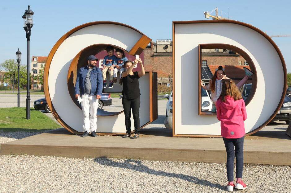 Na zdjęciu widać grupę osób robiących sobie pamiątkowe zdjęcie przy dużym napisie „GDAŃSK”, będącym popularną atrakcją turystyczną. Dzieci i dorośli pozują w literach „G” i „D”, korzystając z ich przestrzennej formy jako ram do zdjęć. 