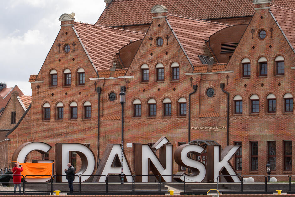 Na zdjęciu widoczny jest duży napis „GDAŃSK” ustawiony na tle ceglanego budynku Polskiej Filharmonii Bałtyckiej na Ołowiance. Przed napisem para turystów robi sobie zdjęcie, co podkreśla popularność tego miejsca jako atrakcji fotograficznej.
