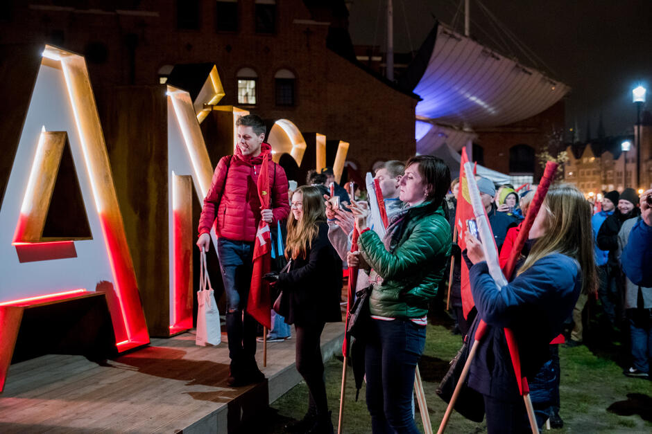 Na zdjęciu widać grupę ludzi zgromadzonych nocą przy podświetlonym napisie „GDAŃSK”, wielu z nich trzyma flagi i robi zdjęcia telefonami. Atmosfera jest radosna i podniosła, sugerując udział w wydarzeniu patriotycznym lub miejskim święcie. 