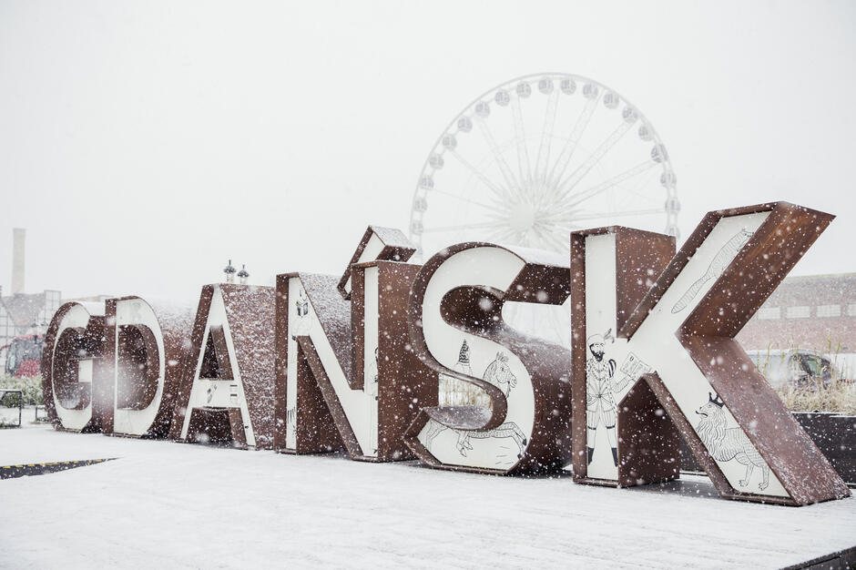 Na zdjęciu przedstawiono ośnieżony napis „GDAŃSK” ozdobiony ilustracjami na tle dużego diabelskiego młyna, co tworzy zimową i bajkową atmosferę. Padający śnieg i biała sceneria podkreślają urok pierwszych dni zimy w mieście.
