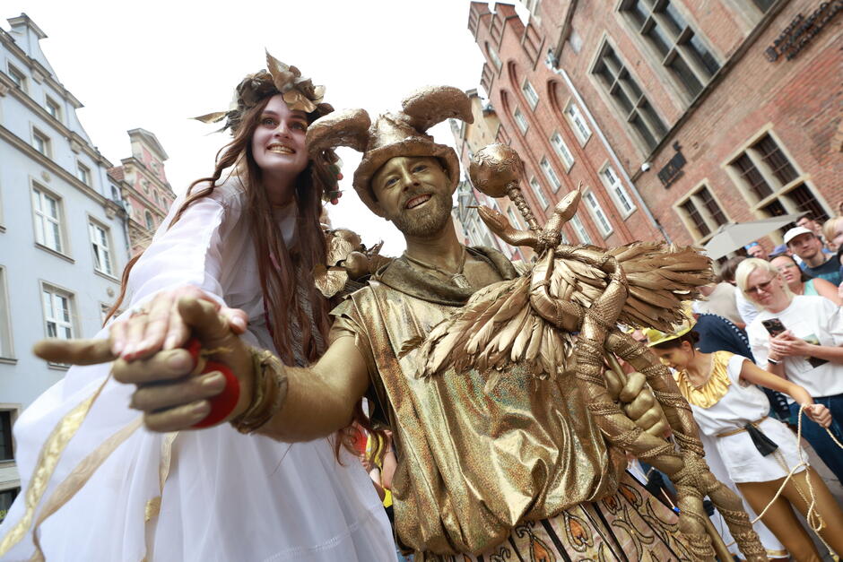 Dwie postaci z uroczystości otwarcia Jarmarku w roku 2024 - pomalowany na złoto aktor w roli Hermesa i towarzysząca aktorka jako jedna z nimf, na biało 
