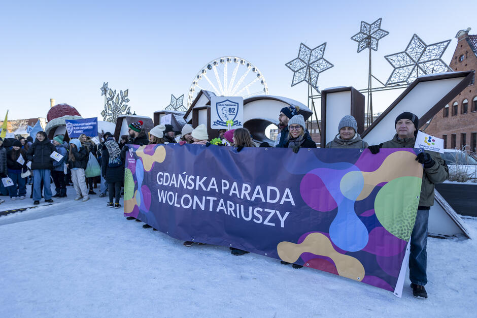 Na tle dużych liter tworzących napis „GDAŃSK” uczestnicy trzymają kolorowy baner z napisem „GDAŃSKA PARADA WOLONTARIUSZY”, uśmiechając się mimo zimowej aury. W tle widoczne są dekoracje świąteczne oraz diabelski młyn, co wskazuje na radosne wydarzenie odbywające się w okresie zimowym.
