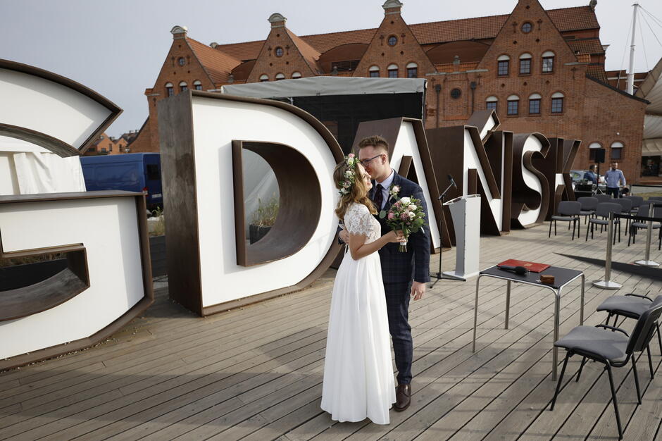 Para młoda całuje się na tle dużych liter tworzących napis „GDAŃSK”, co wskazuje na romantyczną ceremonię ślubną w plenerze. W tle widać charakterystyczne ceglane budynki, a dekoracje i ustawione krzesła sugerują uroczystość zorganizowaną na świeżym powietrzu.
