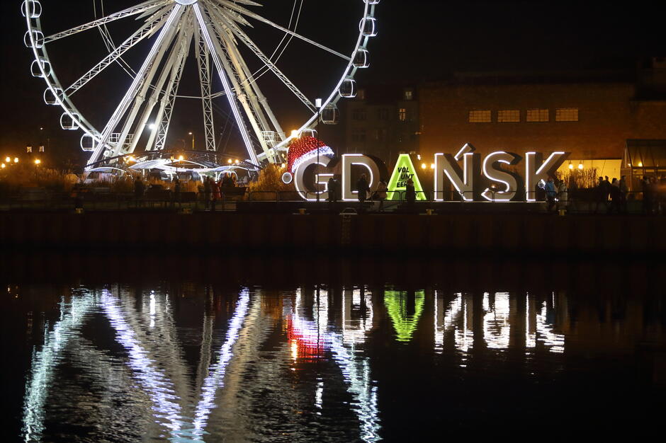 Nocna panorama Gdańska ukazuje podświetlony napis „GDAŃSK” oraz olbrzymi diabelski młyn odbijające się malowniczo w wodzie. Litera „G” ozdobiona jest świąteczną czapką Mikołaja, co nadaje scenerii wyjątkowy, zimowo-świąteczny klimat. A  ma na sobie zieloną tkaninę z napisem Volunteer

