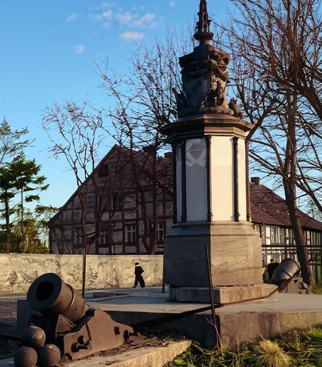 Cokół z pomnikiem, po obu stronach stoją dwa moździerze z tyłu dom szachulcowy