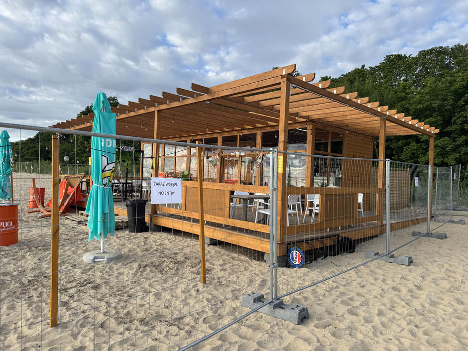 Na zdjęciu widoczna jest zamknięta strefa na plaży z drewnianym barem w tle, otoczona metalowym ogrodzeniem. Przed wejściem umieszczono tabliczkę z napisem „Zakaz wstępu”, a wokół stoją czerwone beczki z logo Soplica i zamknięte parasole turkusowe.