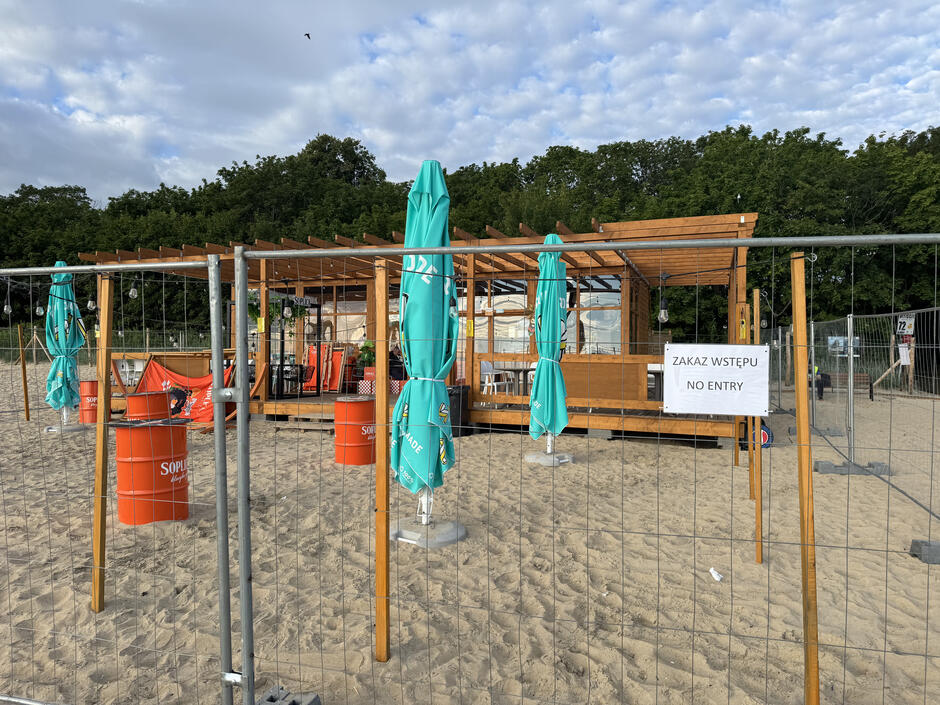 Na zdjęciu widoczna jest zamknięta strefa na plaży z drewnianym barem w tle, otoczona metalowym ogrodzeniem. Przed wejściem umieszczono tabliczkę z napisem „Zakaz wstępu”, a wokół stoją czerwone beczki z logo Soplica i zamknięte parasole turkusowe.