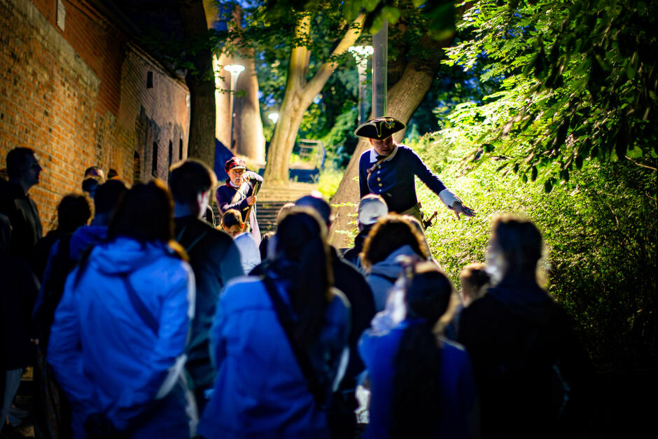 Na zdjęciu widoczna jest grupa ludzi uczestnicząca w nocnym spacerze z przewodnikiem w historycznym stroju, który opowiada historię lub legendę, wskazując coś w zaroślach. W tle widać drugiego aktora w kostiumie z muszkietem, co nadaje wydarzeniu teatralny i edukacyjny charakter.
