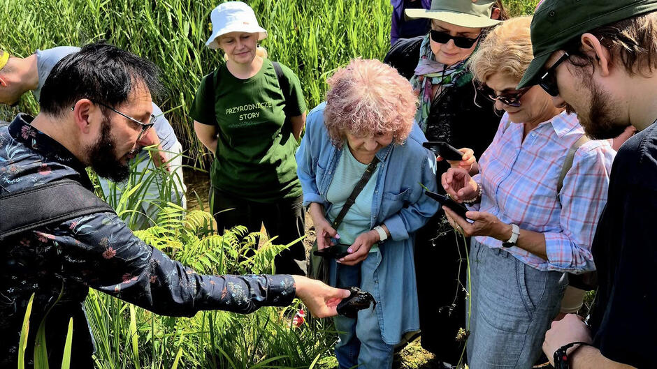 Grupa osób obserwuje z bliska okazałego czarne zwierze trzymane przez mężczyznę wśród wysokiej trawy. Część z nich robi zdjęcia telefonami, a w tle widać zielone szuwary.