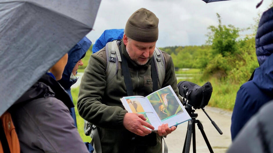 Mężczyzna w czapce i kurtce pokazuje książkę z ilustracjami ptaków, tłumacząc coś grupie osób ubranych na deszczową pogodę. Obok stoi statyw z lunetą, a w tle widać polną drogę i zielone krzewy.