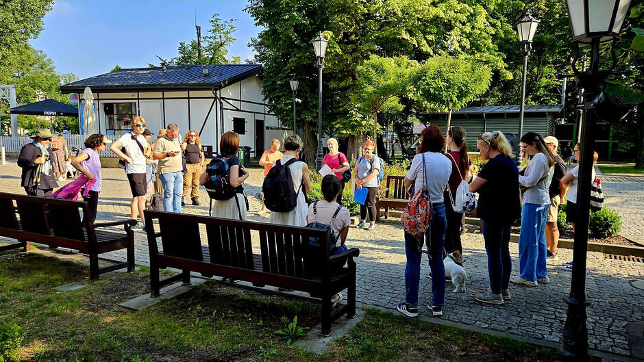 Grupa ludzi zgromadzona na brukowanej alejce w parku, słucha osoby prowadzącej spotkanie lub wycieczkę. Wokół znajdują się ławki, latarnie i drzewa, a towarzyszy im słoneczna pogoda.