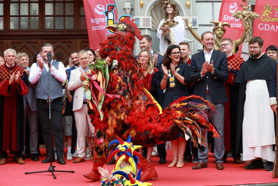 Przed grupą oficjeli i widzów stoi ogromny kolorowy kogut – postać w kostiumie z piór i wstążek – będąca centralnym punktem ceremonii. W tle widać flagi z logo Gdańska oraz przedstawicieli władz i organizatorów, którzy oklaskują występ.