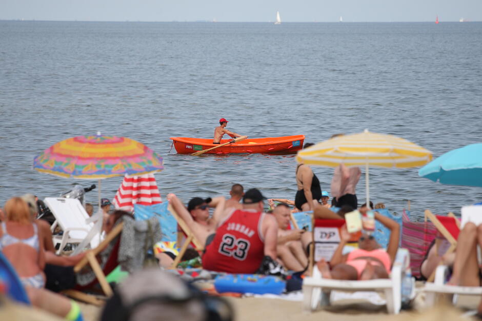 Tłoczna plaża z kolorowymi parasolami i leżakami, na której wypoczywa wiele osób. W tle na wodzie widać ratownika w czerwonej czapce, wiosłującego w pomarańczowej łodzi.