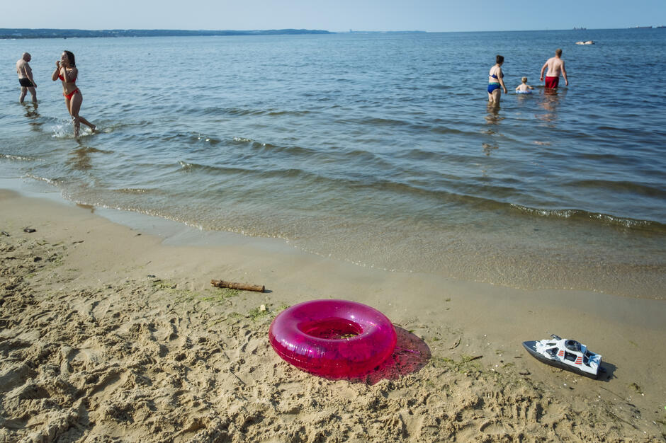 Spokojna plaża z kilkoma osobami kąpiącymi się w morzu, a na brzegu leży różowe dmuchane koło i zabawkowa motorówka. W tle widać horyzont i zarys lądu po drugiej stronie zatoki.
