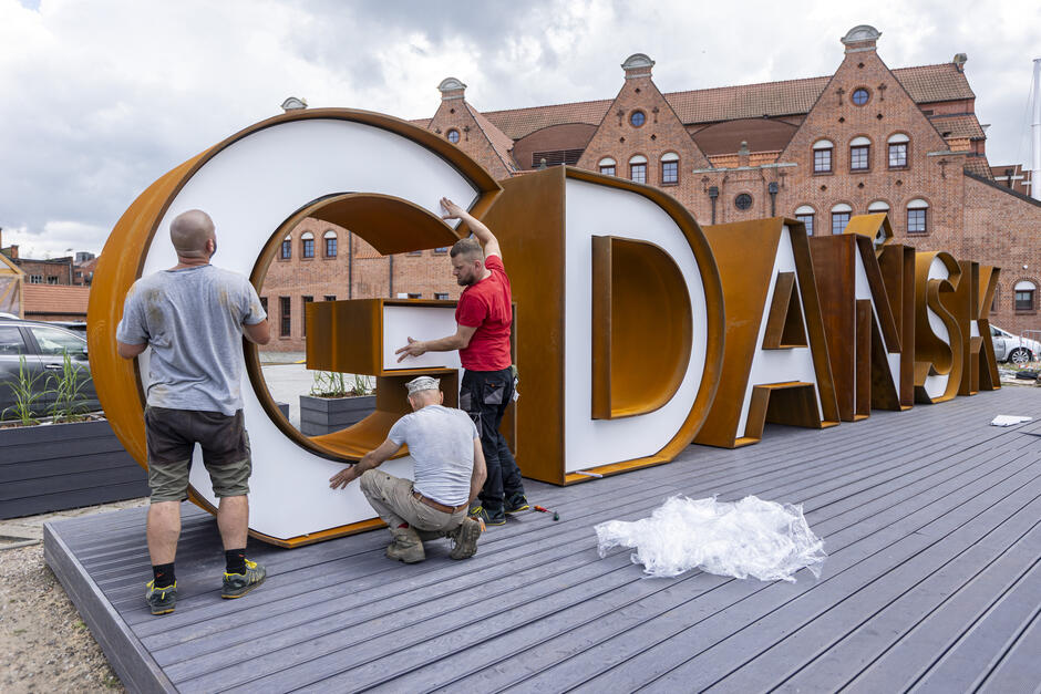 Na zdjęciu widać trzech mężczyzn montujących duży, przestrzenny napis „GDAŃSK” na nabrzeżu w pobliżu zabytkowych budynków z czerwonej cegły. Litery są nowoczesne, w kolorze rdzawego metalu z białym wypełnieniem, a całość powstaje na drewnianym podeście.
