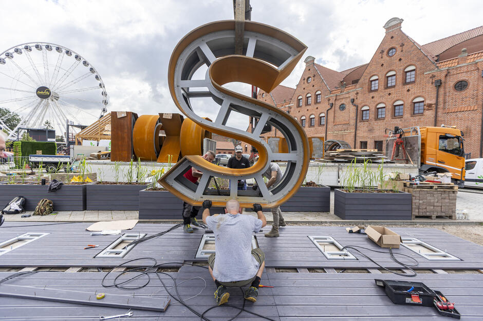 Na zdjęciu kilku pracowników montuje ogromną literę „S” należącą do napisu „GDAŃSK” na placu w okolicach Ołowianki. W tle widać charakterystyczny budynek z czerwonej cegły oraz diabelski młyn, co wskazuje na turystyczną i miejską scenerię. 
