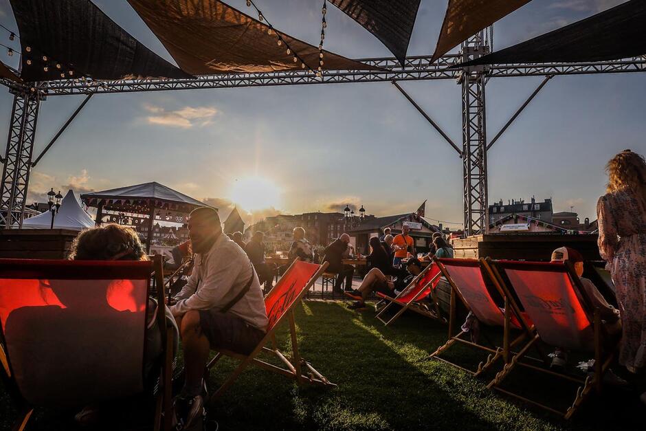 Na zdjęciu widać relaksującą strefę wypoczynku z leżakami, gdzie ludzie spędzają czas przy zachodzącym słońcu podczas letniego wydarzenia plenerowego. Przestrzeń jest zadaszona lekką konstrukcją z materiału, a wokół panuje swobodna, festiwalowa atmosfera.
