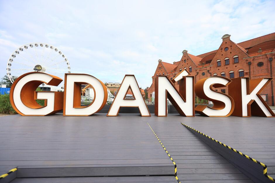 Na zdjęciu widnieje duży, przestrzenny napis „GDAŃSK” ustawiony na drewnianym podeście, z charakterystyczną zabytkową ceglaną architekturą w tle. Po lewej stronie widać diabelski młyn, który dodaje nowoczesnego akcentu tej historycznej przestrzeni.
