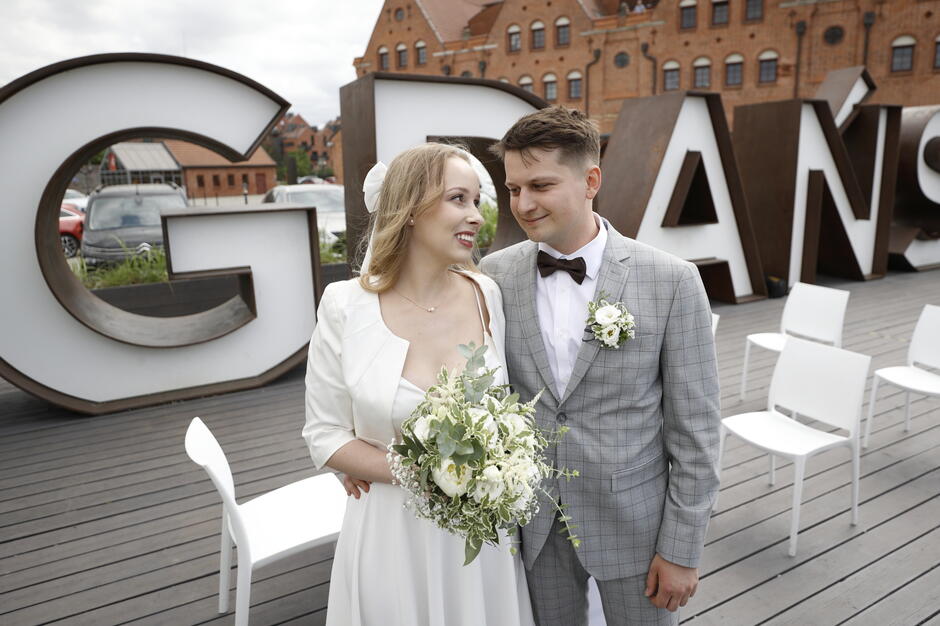 Na zdjęciu widzimy uśmiechniętą parę młodą podczas ślubu plenerowego, stojącą na tle dużego napisu „GDAŃSK” i zabytkowych, ceglanych budynków. Panna młoda trzyma elegancki bukiet z białych kwiatów, a pan młody ubrany jest w jasnoszary garnitur w kratę z muchą i przypiętą butonierką. 