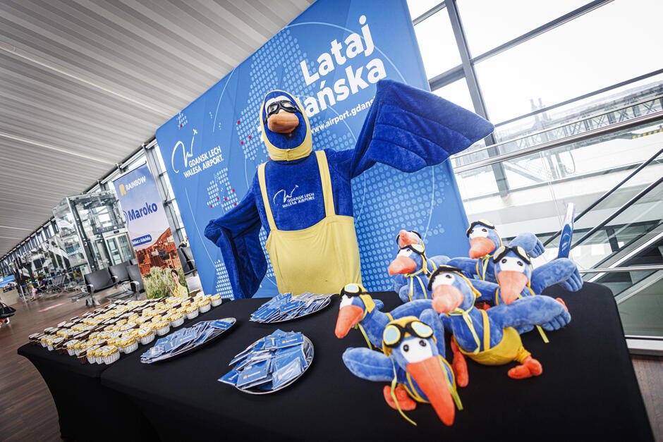 Na zdjęciu widzimy promocyjne stoisko Portu Lotniczego Gdańsk im. Lecha Wałęsy z maskotką w kształcie niebieskiego ptaka w lotniczym stroju oraz pluszowymi wersjami tej postaci. Na stole znajdują się także przekąski i materiały promocyjne, a całość zaaranżowano w przestrzeni lotniskowej.
