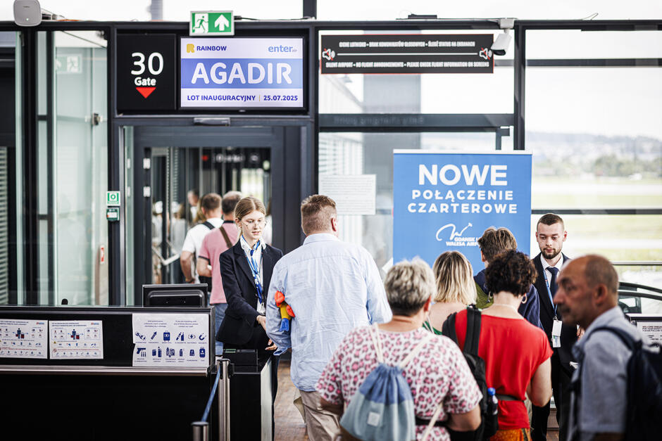Na zdjęciu pasażerowie ustawiają się w kolejce do bramki 30 na gdańskim lotnisku, przygotowując się do inaugurującego lotu czarterowego do Agadiru, który odbywa się 25 lipca 2025 roku. Obok bramki znajduje się baner informujący o nowym połączeniu czarterowym zorganizowanym przez Rainbow i Enter Air.
