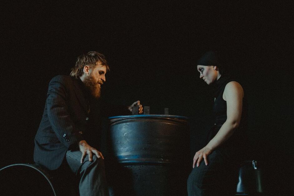 Dwoje aktorów siedzi naprzeciwko siebie przy metalowej beczce, w półmroku, z poważnymi minami. Scena ma mroczny, teatralny klimat.